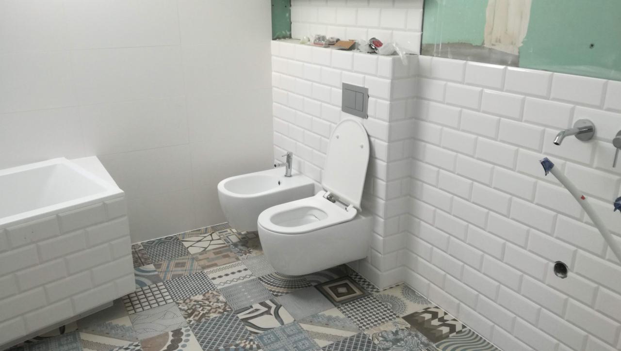 Biała łazienka w trakcie wykańczania: białe płytki typu metro na ścianach, patchworkowa podłoga, wisząca toaleta i bidet, widoczne elementy instalacji wodnej, surowy stan wykończenia w górnej...