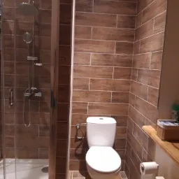 Wykończona łazienka z kabiną prysznicową i toaletą, wyłożona brązowymi płytkami imitującymi drewno, z widocznym grzejnikiem i uchwytem na papier toaletowy.