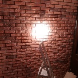 Robert Krzemiński - Ściana wyłożona dekoracyjną cegłą w odcieniach czerwieni i brązu, z widoczną drabiną i materiałami budowlanymi sugerującymi etap wykończeniowy.