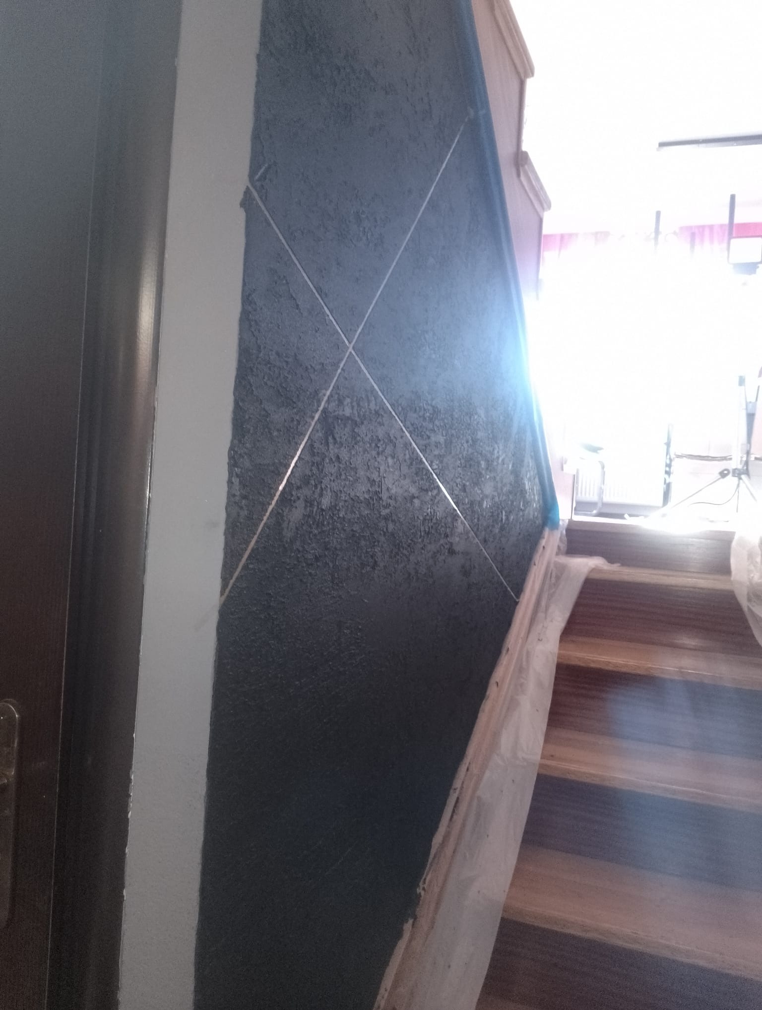 Ciemna, teksturowana ściana z dekoracyjnymi, metalicznymi paskami ułożonymi w kształt rombów, sąsiadująca z pomalowaną na biało ścianą przy drzwiach, z zabezpieczonymi folią schodami w tle.