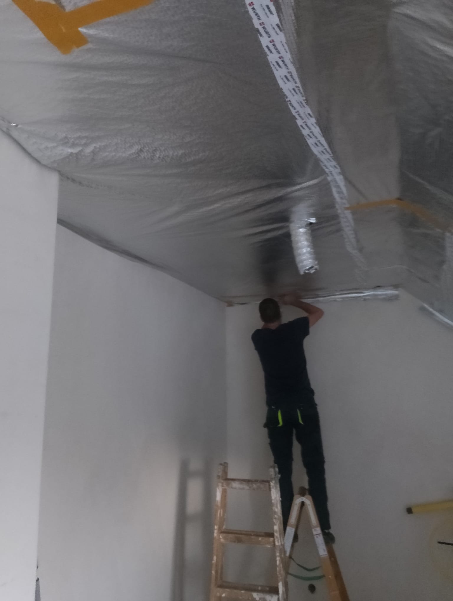 Pracownik na drabinie montuje izolację z folii aluminiowej na skośnym suficie poddasza, zabezpieczoną taśmą marki Wurth.