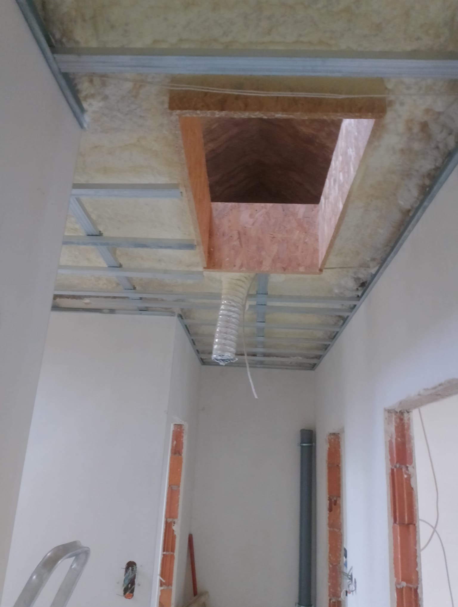 Sufit podwieszany w trakcie montażu z izolacją z wełny mineralnej, widoczne profile stalowe, otwór wentylacyjny z podłączonym przewodem elastycznym i częściowo wykończone ściany z ościeżnicami.