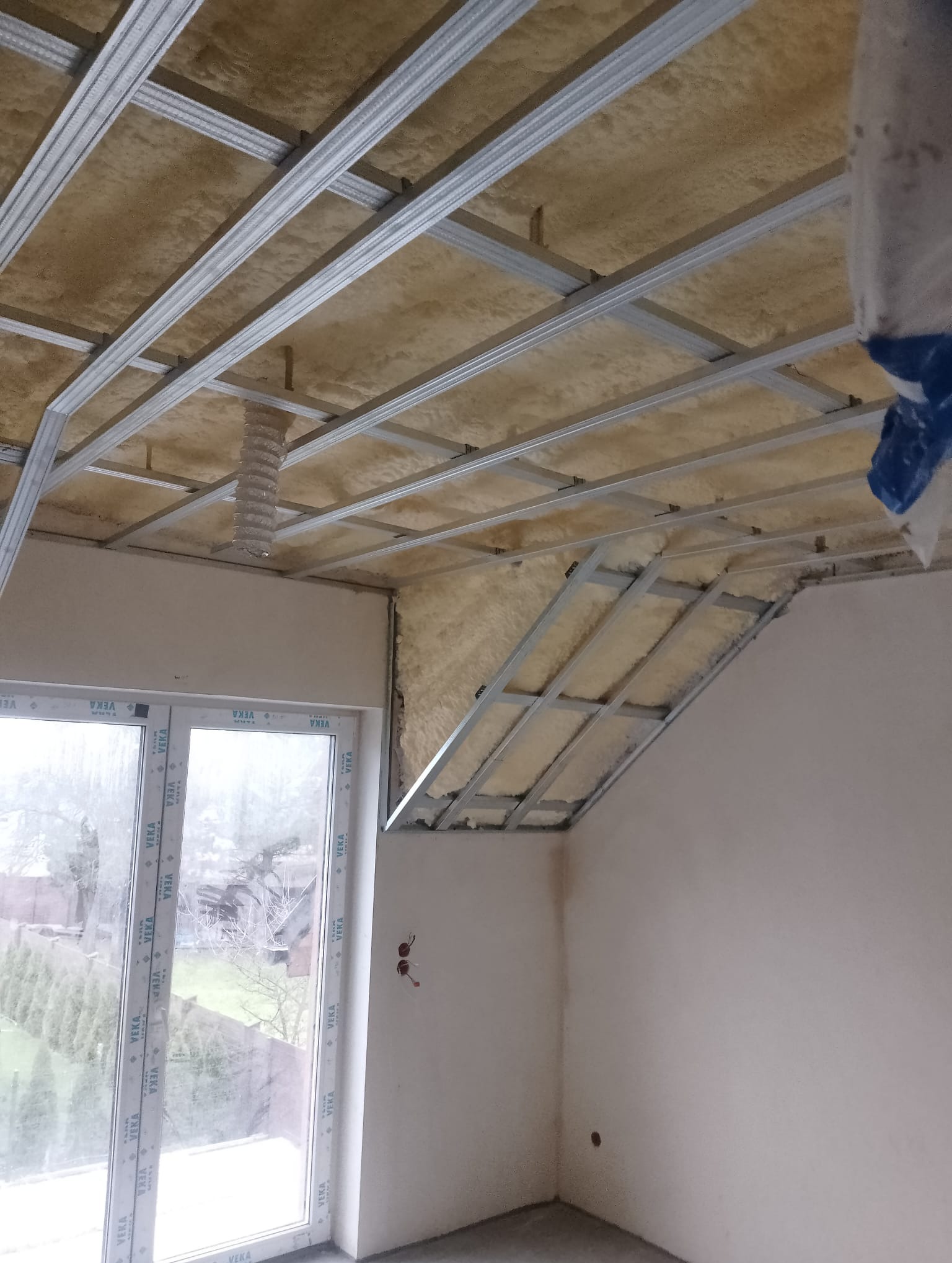 Montaż stelaża sufitu podwieszanego z izolacją w Pszczynie, widoczne profile metalowe i wełna mineralna, okno z widokiem na zewnątrz.