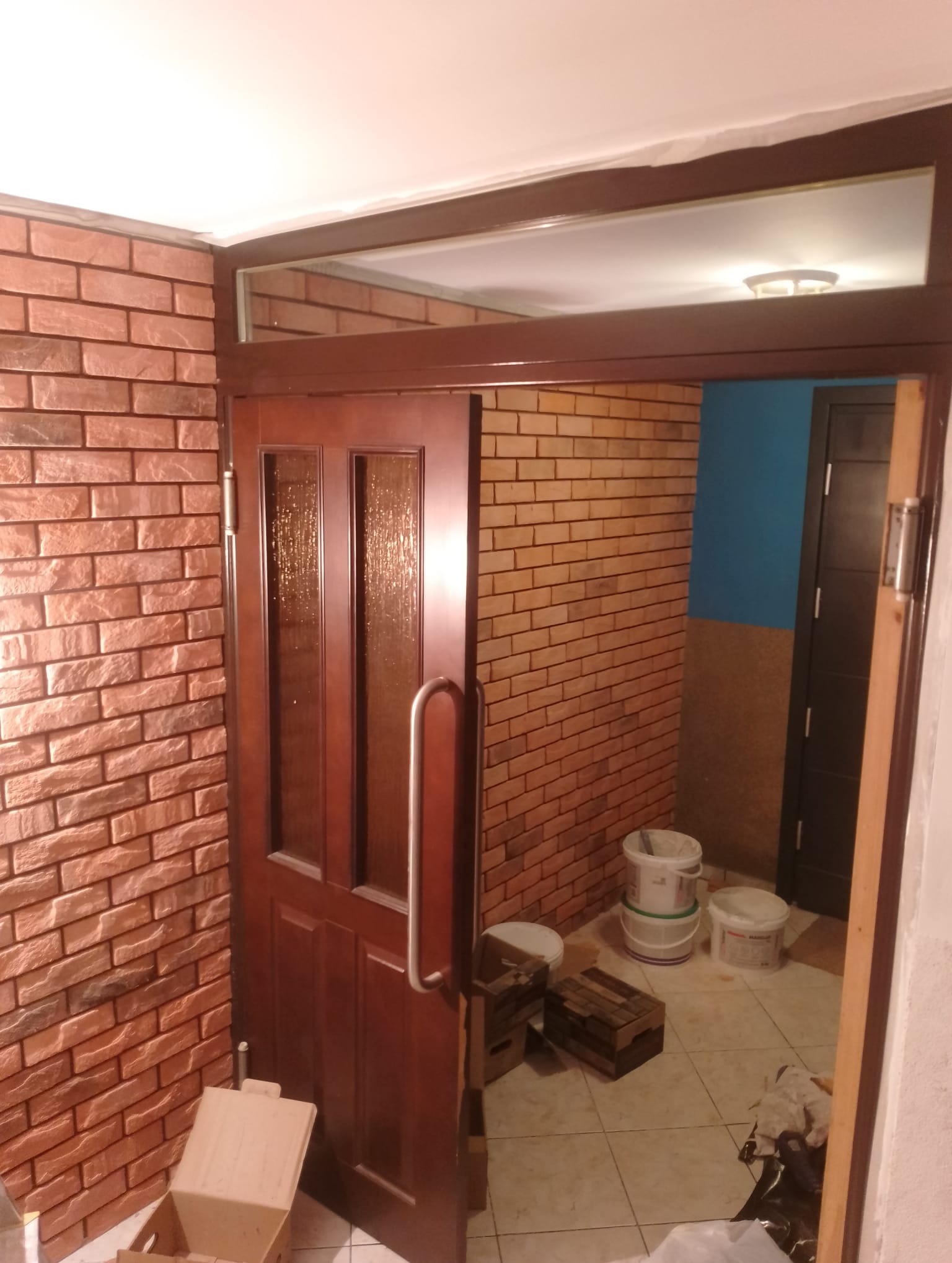 Korytarz z ceglaną ścianą, drewnianymi drzwiami z szybą i wiadrami z farbą na podłodze z płytek.