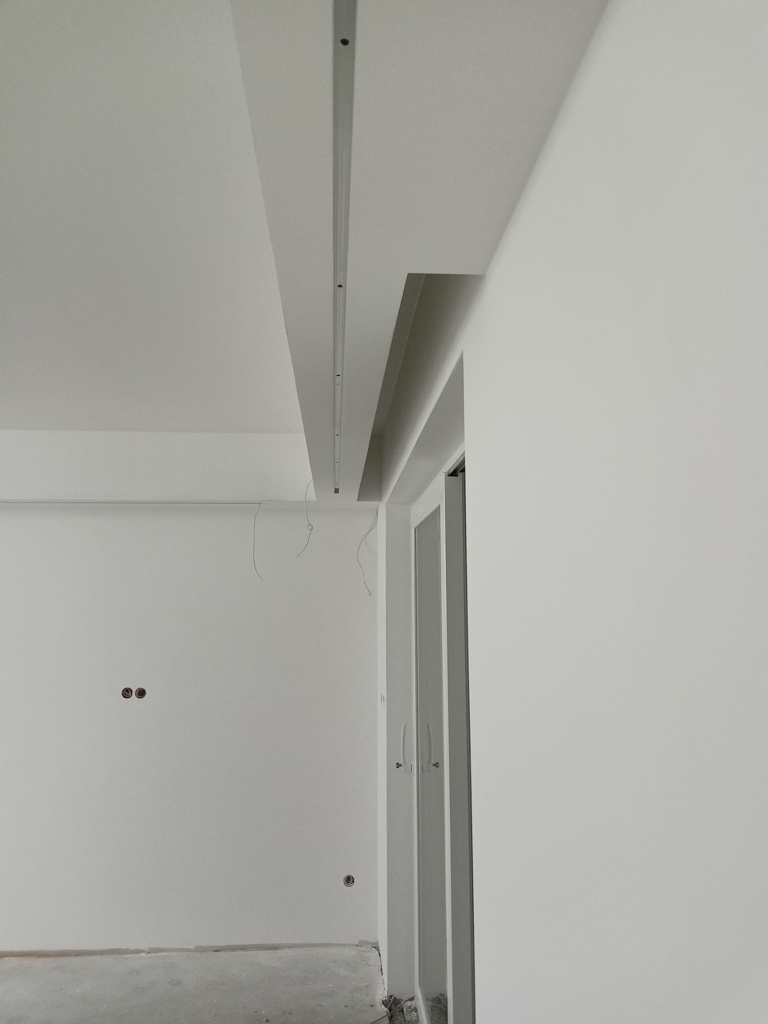 Wnętrze w trakcie prac wykończeniowych: ściany pomalowane na biało, widoczne puszki elektryczne, zamontowany profil oświetleniowy na suficie podwieszanym, drzwi wejściowe.