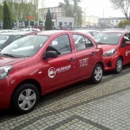 Czerwone samochody marki Nissan i Toyota z logo firmy Helpacar oferującej auto zastępcze z OC sprawcy, zaparkowane na kostce brukowej przed budynkiem.