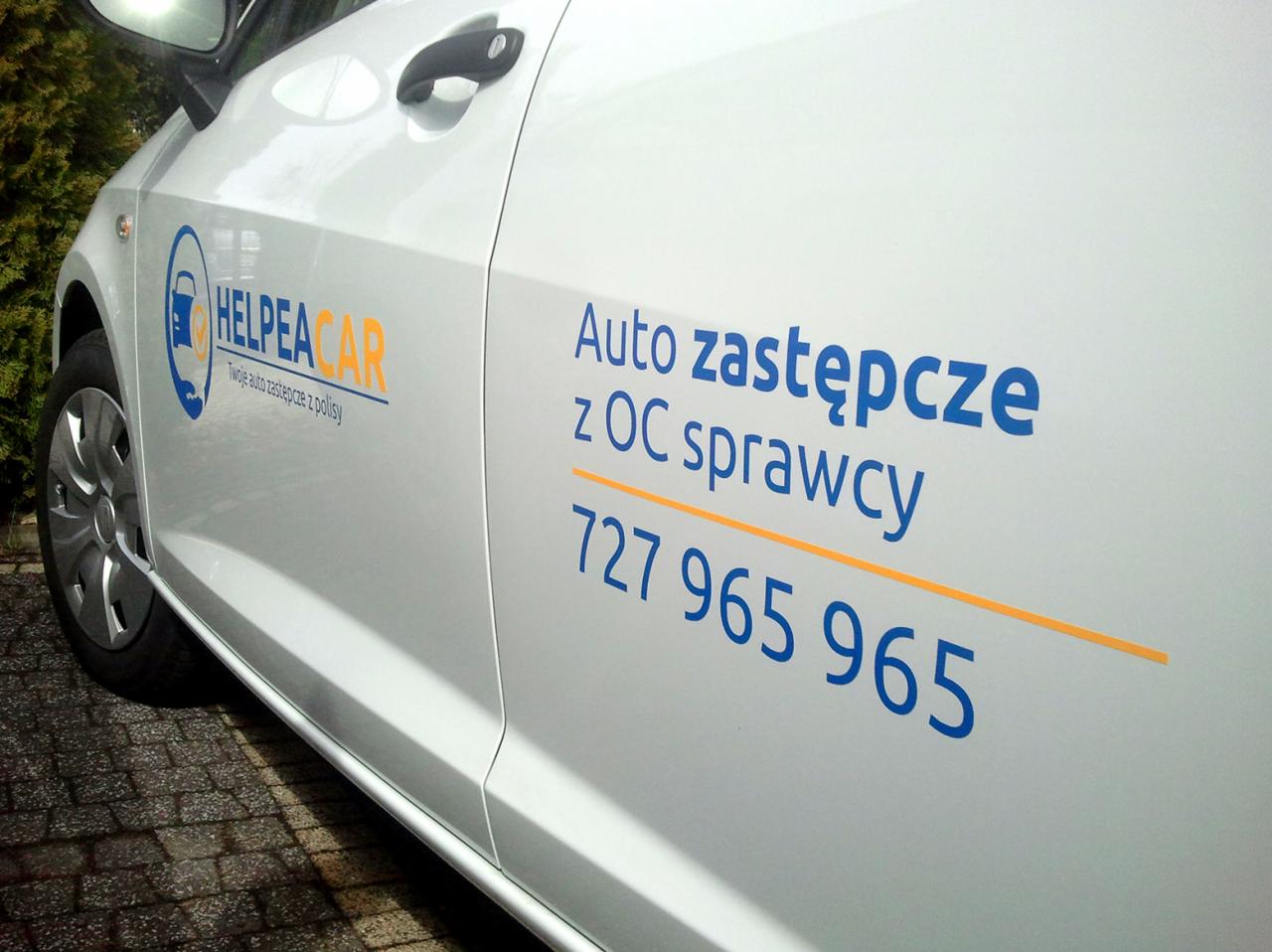 Biały samochód z logo firmy Helpea Car i napisem 'Auto zastępcze z OC sprawcy' oraz numerem telefonu, zaparkowany na kostce brukowej.