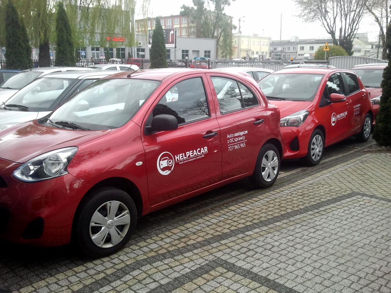 Czerwone samochody marki Nissan i Toyota z logo firmy Helpacar oferującej auto zastępcze z OC sprawcy, zaparkowane na kostce brukowej przed budynkiem.