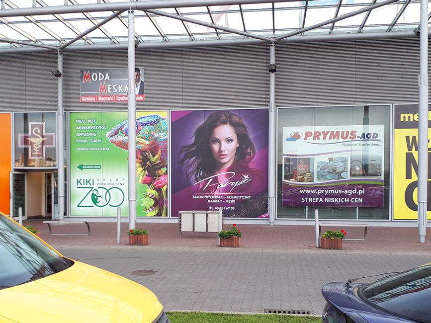 Zewnętrzny widok na budynek z wielkoformatowymi reklamami różnych firm: salonu fryzjersko-kosmetycznego, sklepu zoologicznego, sklepu z odzieżą męską i sklepu AGD. Widoczne zaparkowane samochody.