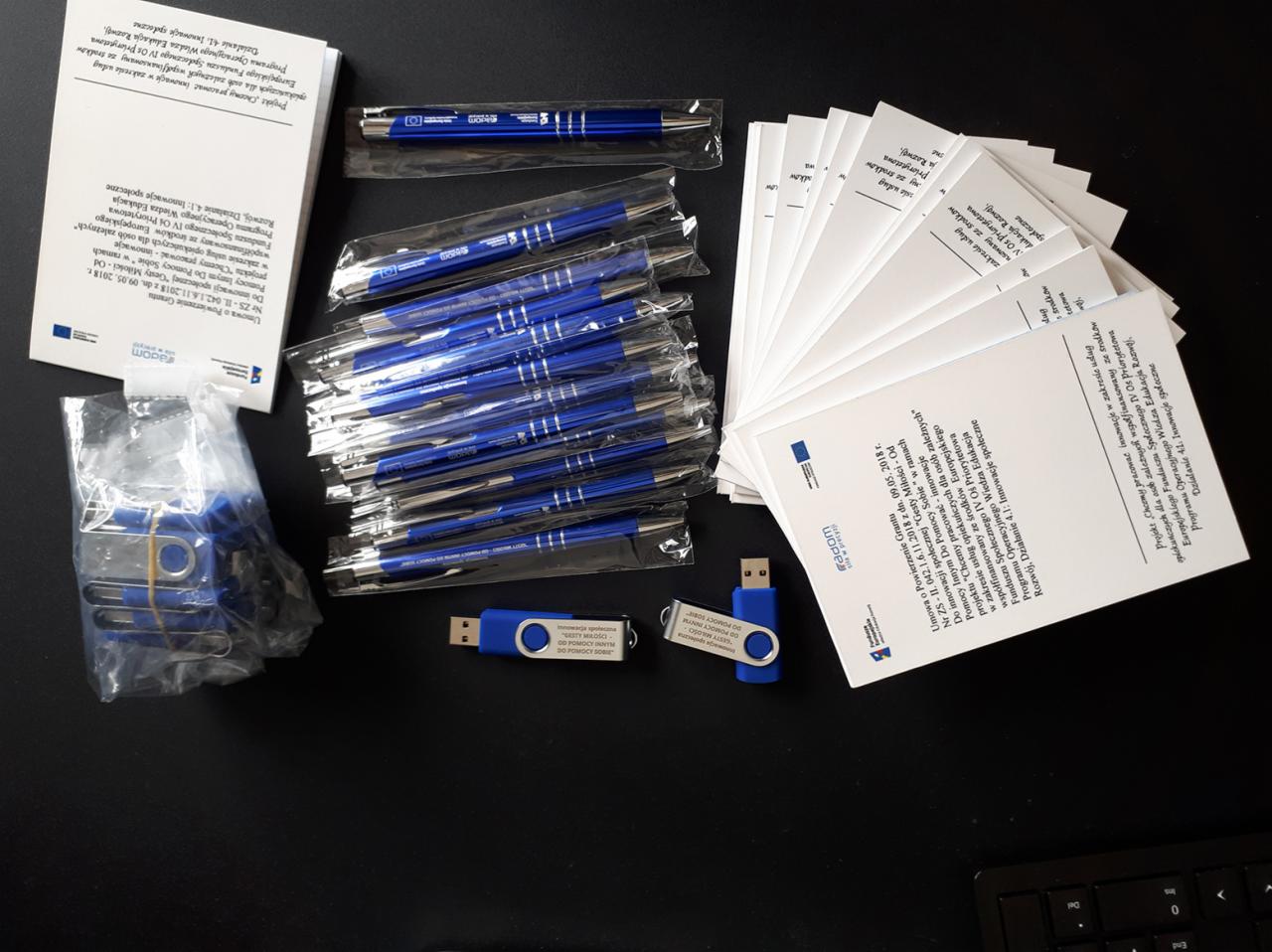 Zestaw promocyjny z niebieskimi długopisami w folii, pendrive'ami z logo i ulotkami informacyjnymi na ciemnym tle biurka.