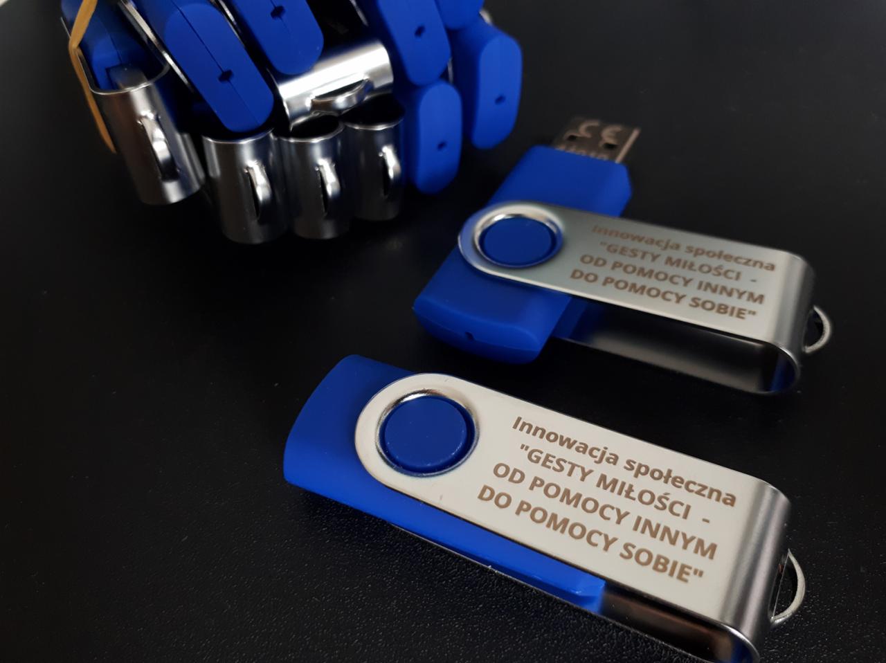 Niebieskie pendrive'y z metalową obudową, na której wygrawerowano napis 'Innowacja społeczna 'GESTY MIŁOŚCI - OD POMOCY INNYM DO POMOCY SOBIE'', leżące na czarnym tle obok mechanicznej ręki...