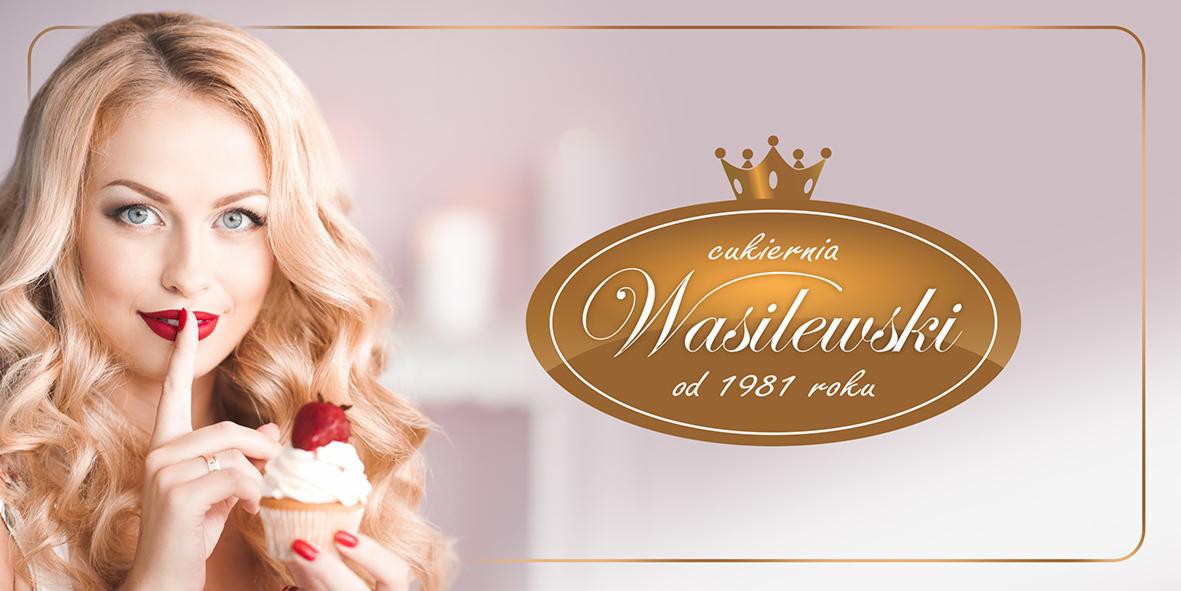 Blondwłosa kobieta z czerwonymi ustami trzyma babeczkę z truskawką, palcem wskazującym przy ustach, obok logo cukierni Wasilewski z 1981 roku na rozmytym tle.
