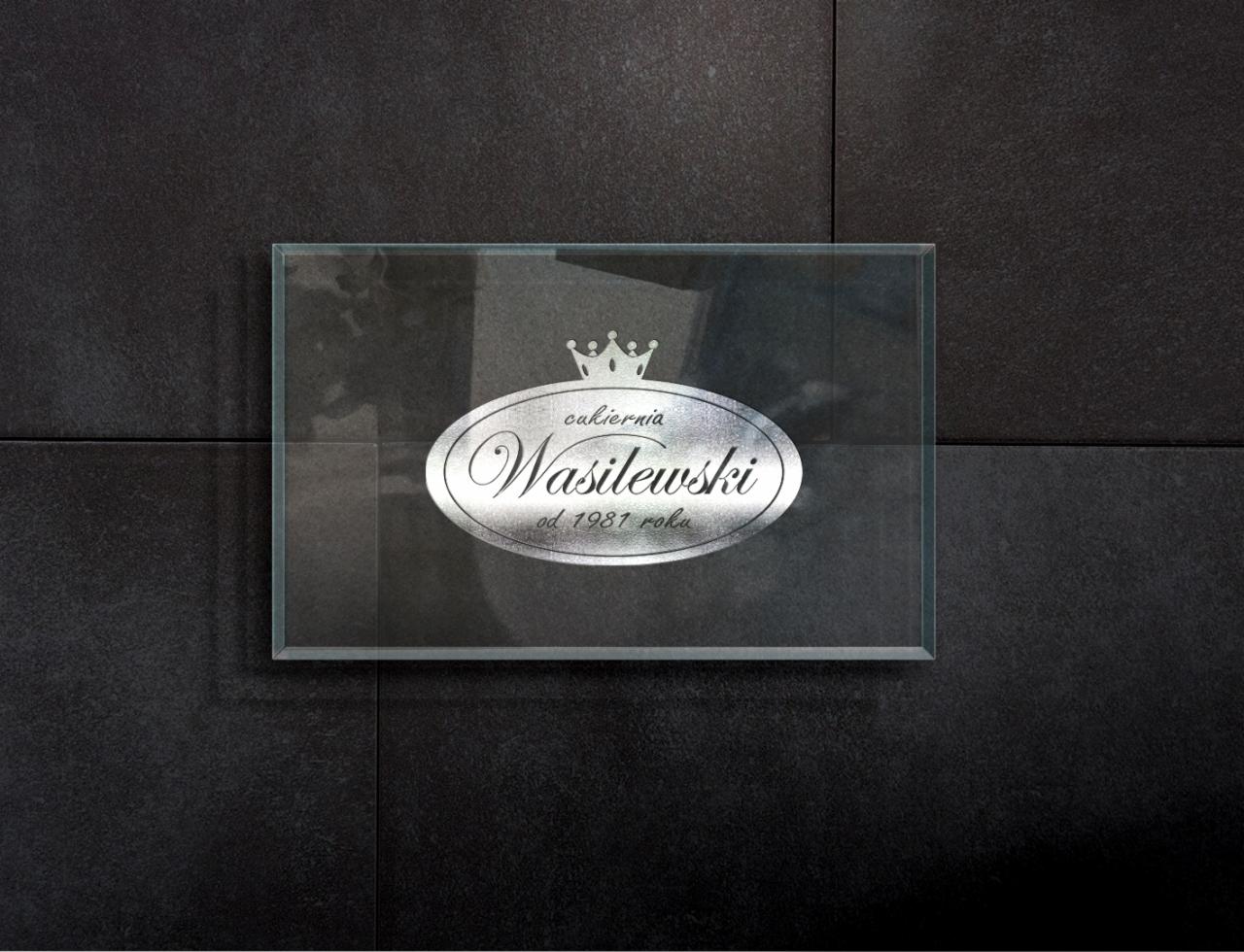 Szklana tabliczka z logo cukierni Wasilewski, umieszczona na ciemnej, strukturalnej ścianie z płytek.