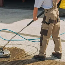 Pracownik w kombinezonie roboczym czyści kostkę brukową za pomocą myjki ciśnieniowej z okrągłą szczotką, w tle widoczna ż&oacute;łta obudowa urządzenia.
