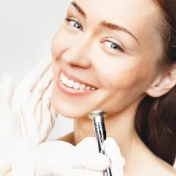 Uśmiechnięta kobieta podczas zabiegu mikrodermabrazji diamentowej, widoczne rękawice ochronne i urządzenie do peelingu na tle jasnego studia.