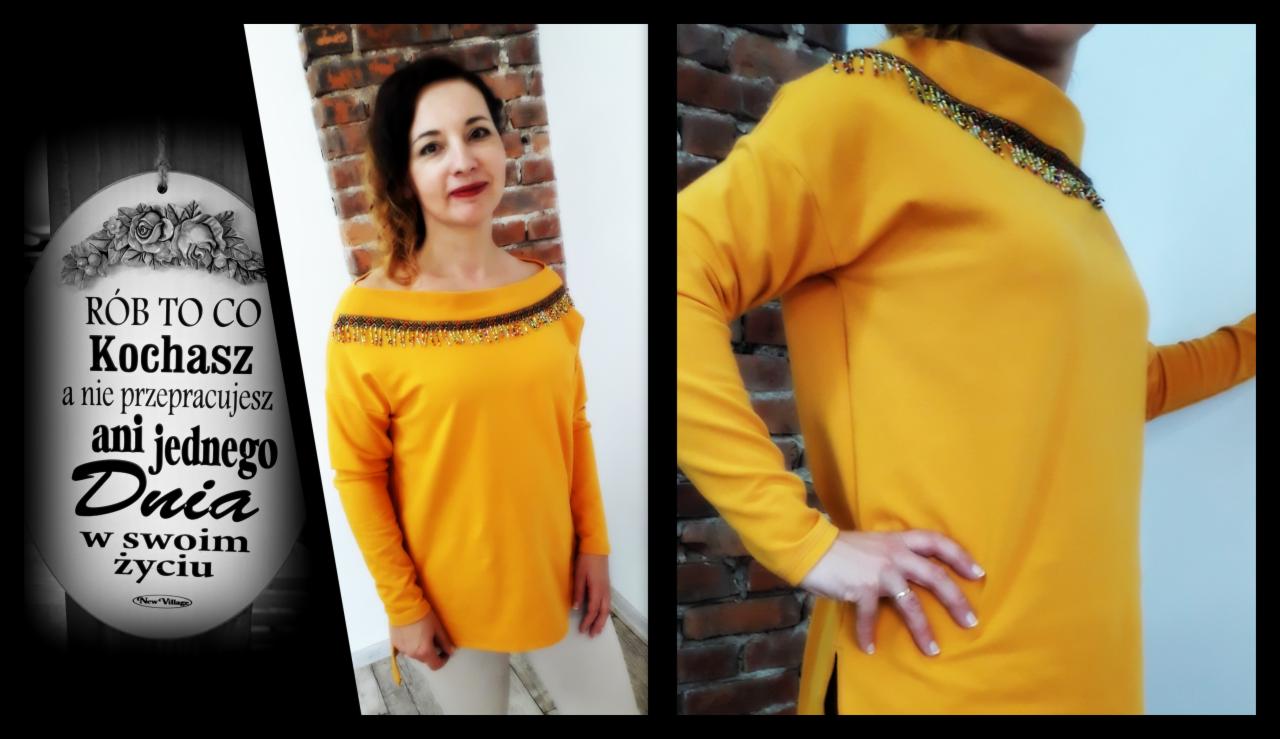 Kobieta w musztardowej bluzce z ozdobnym dekoltem z koralików, prezentacja modelu, na drugim planie ceglana ściana i tabliczka z napisem.