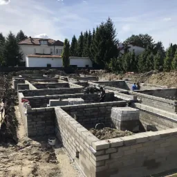 Wykopane i wymurowane z bloczków betonowych fundamenty pod budynek mieszkalny, widoczni pracownicy w trakcie prac murarskich, w tle dom i drzewa.