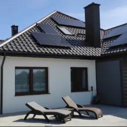 Nowoczesny dom jednorodzinny z czarną dachówką i panelami słonecznymi na dachu, widok na taras z leżakami i pergolą w słoneczny dzień.