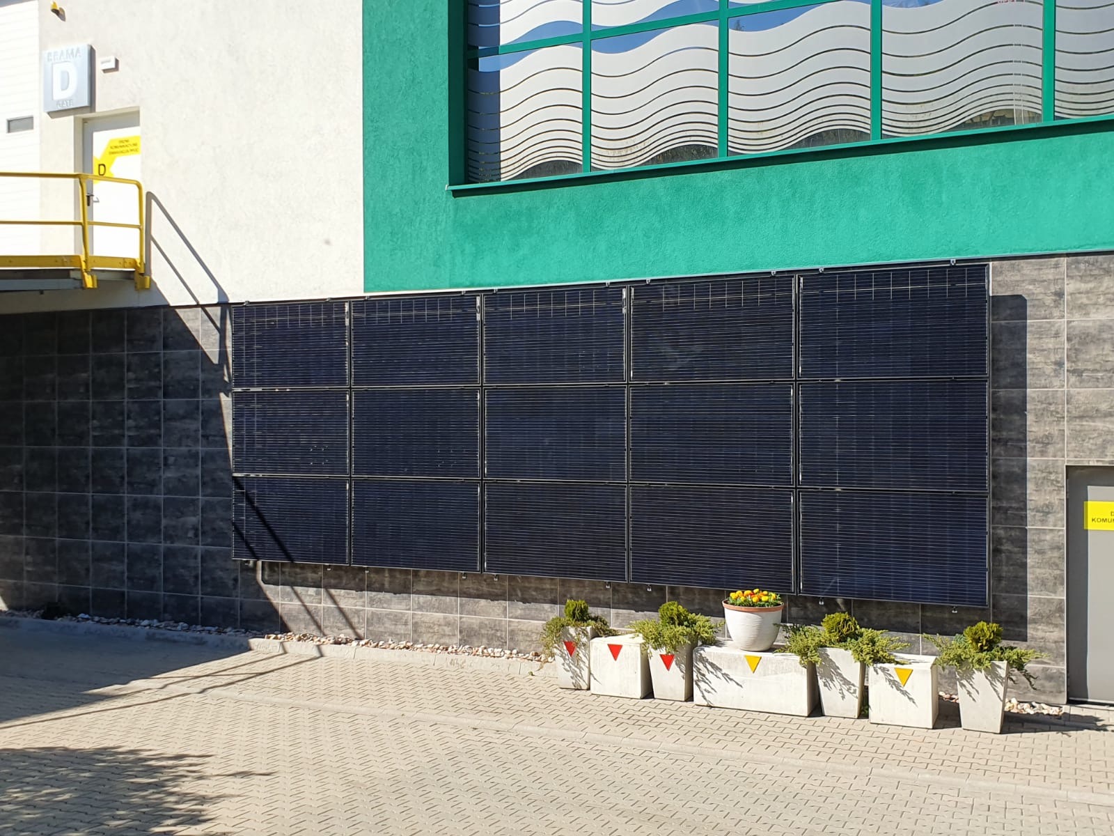 Panele słoneczne zamontowane na szarej ścianie budynku z zielonym akcentem, poniżej rzędu okien z falistym wzorem, ozdobione donicami z roślinami i kwiatami.