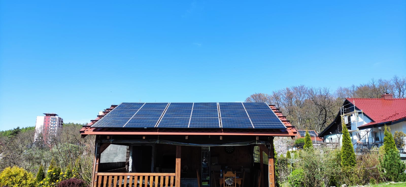 Panele słoneczne na dachu drewnianej altany w ogrodzie, z budynkiem mieszkalnym w tle, błękitne niebo.