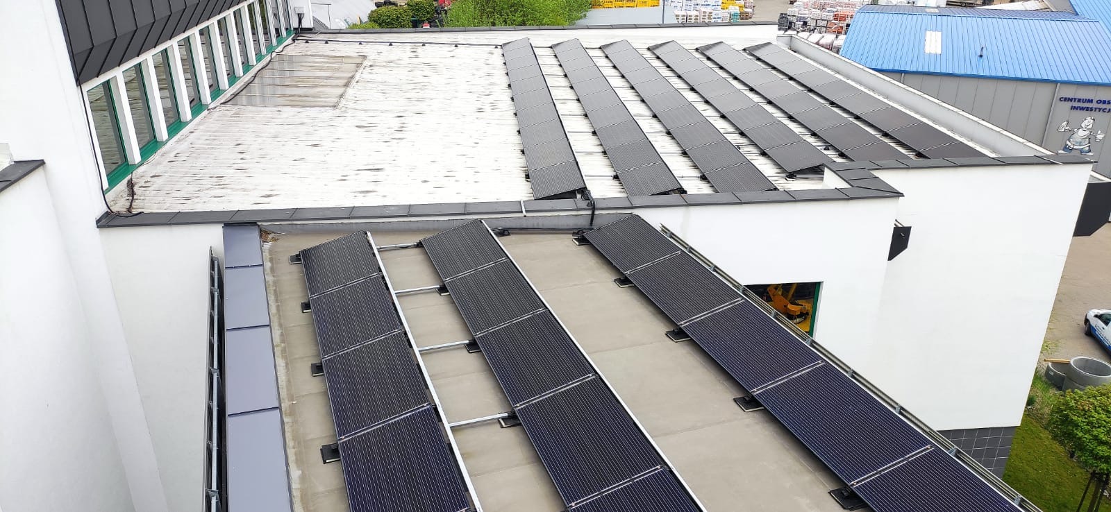 Rząd paneli słonecznych na płaskim dachu budynku komercyjnego, widok z góry pod kątem, z widoczną konstrukcją wsporczą i fragmentem elewacji budynku obok.