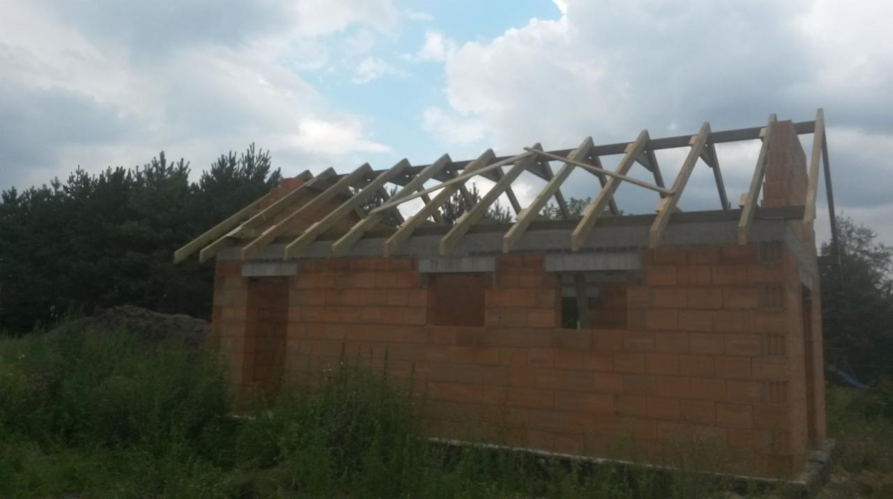 Surowy budynek w trakcie budowy dachu, widoczne drewniane krokwie i mur z pustaków, tło stanowią drzewa i pochmurne niebo.