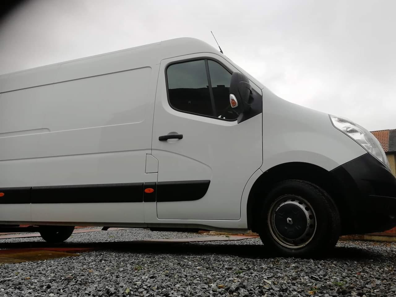 Biały samochód dostawczy Renault Master na żwirowym podłożu, z widocznym fragmentem budynku w tle i pochmurnym niebem.