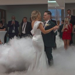 MW Airfilm - Pierwszy taniec pary młodej w otoczeniu dymu, goście obserwują i filmują. Panna młoda w białej sukni, pan młody w ciemnozielonym garniturze. Dekoracja sali.