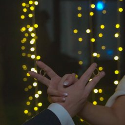 MW Airfilm - Obrączki ślubne na palcach pary młodej w Krakowie, na tle dekoracji świetlnych. Ujęcie z bliska, skupione na dłoniach i symbolice małżeństwa.