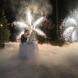 MW Airfilm - Para młoda kroi tort weselny w otoczeniu sztucznych ogni i ciężkiego dymu na tle ciemnego lasu. Na lewo widoczny mężczyzna obsługujący wytwornicę dymu.
