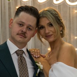 MW Airfilm - Portret pary młodej w zbliżeniu, on w ciemnozielonym garniturze, ona w białej sukni, na tle białej zasłony z delikatnymi światełkami, z subtelnym uśmiechem.