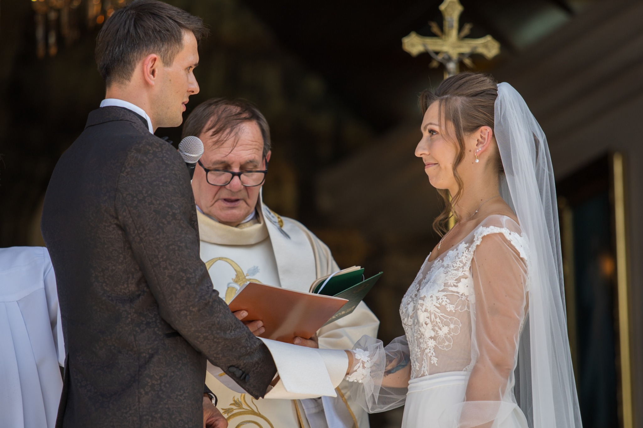 Uśmiechnięta panna młoda w koronkowej sukni i welonie podczas ceremonii ślubnej w kościele, obok stoi pan młody i ksiądz z księgą.