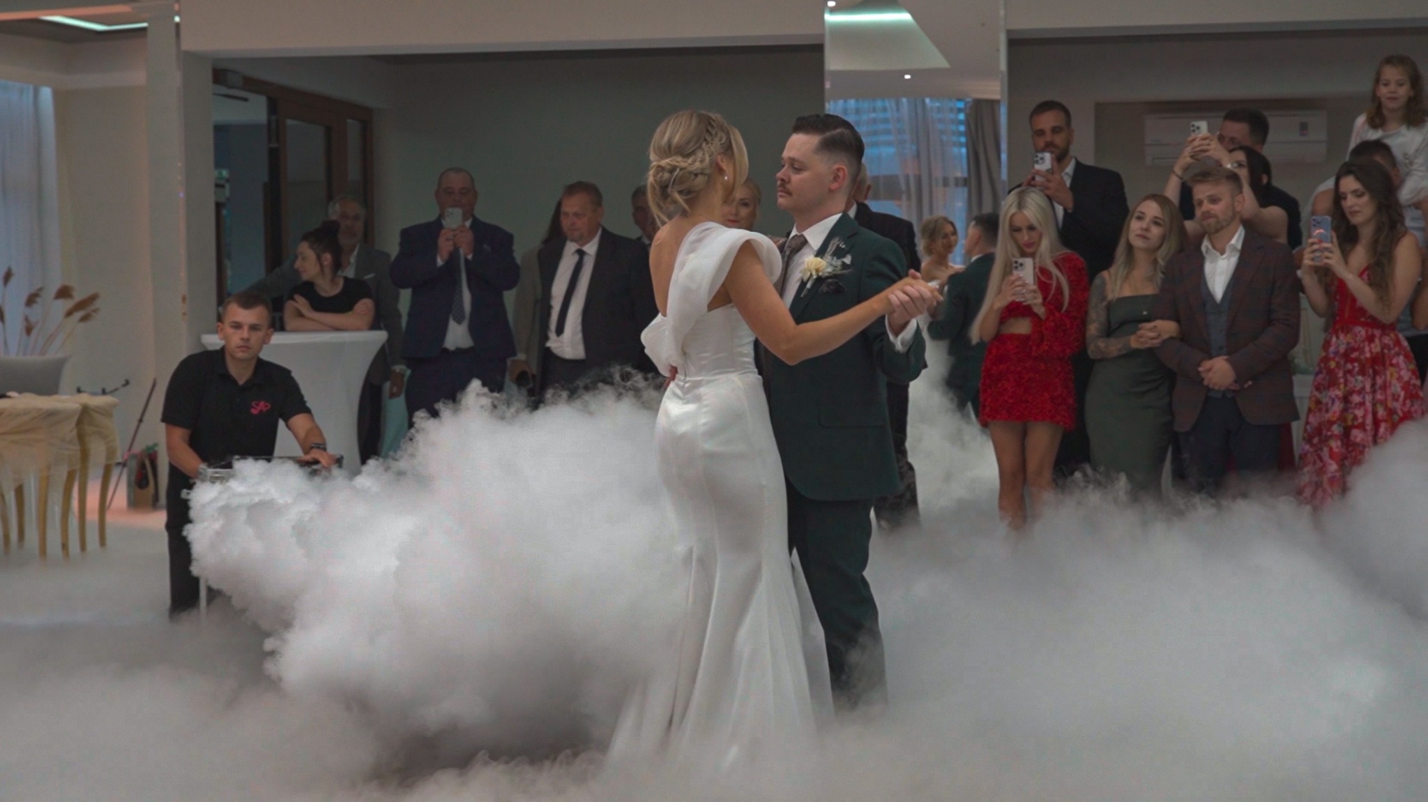 Pierwszy taniec pary młodej w otoczeniu dymu, goście obserwują i filmują. Panna młoda w białej sukni, pan młody w ciemnozielonym garniturze. Dekoracja sali.