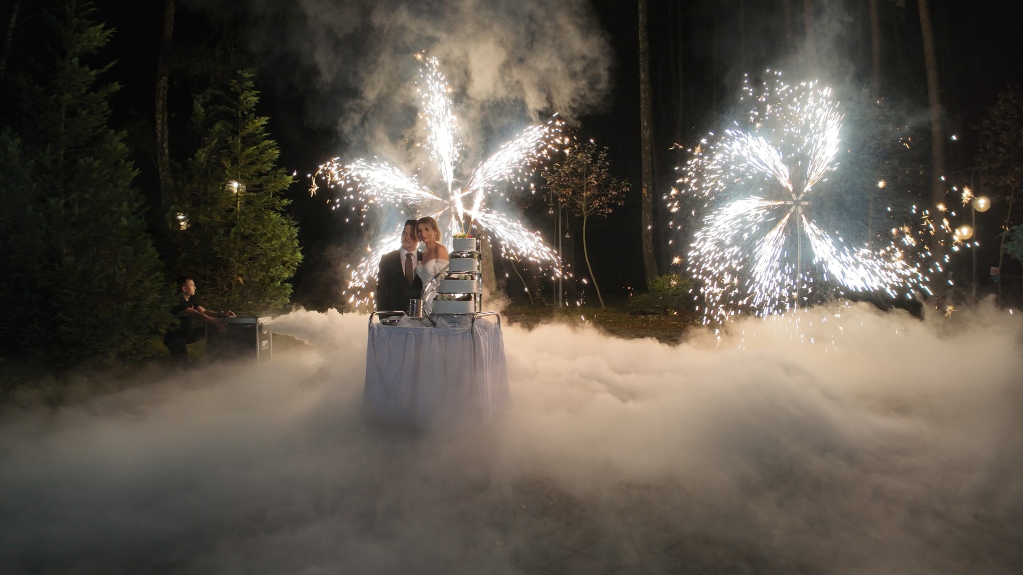 Para młoda kroi tort weselny w otoczeniu sztucznych ogni i ciężkiego dymu na tle ciemnego lasu. Na lewo widoczny mężczyzna obsługujący wytwornicę dymu.