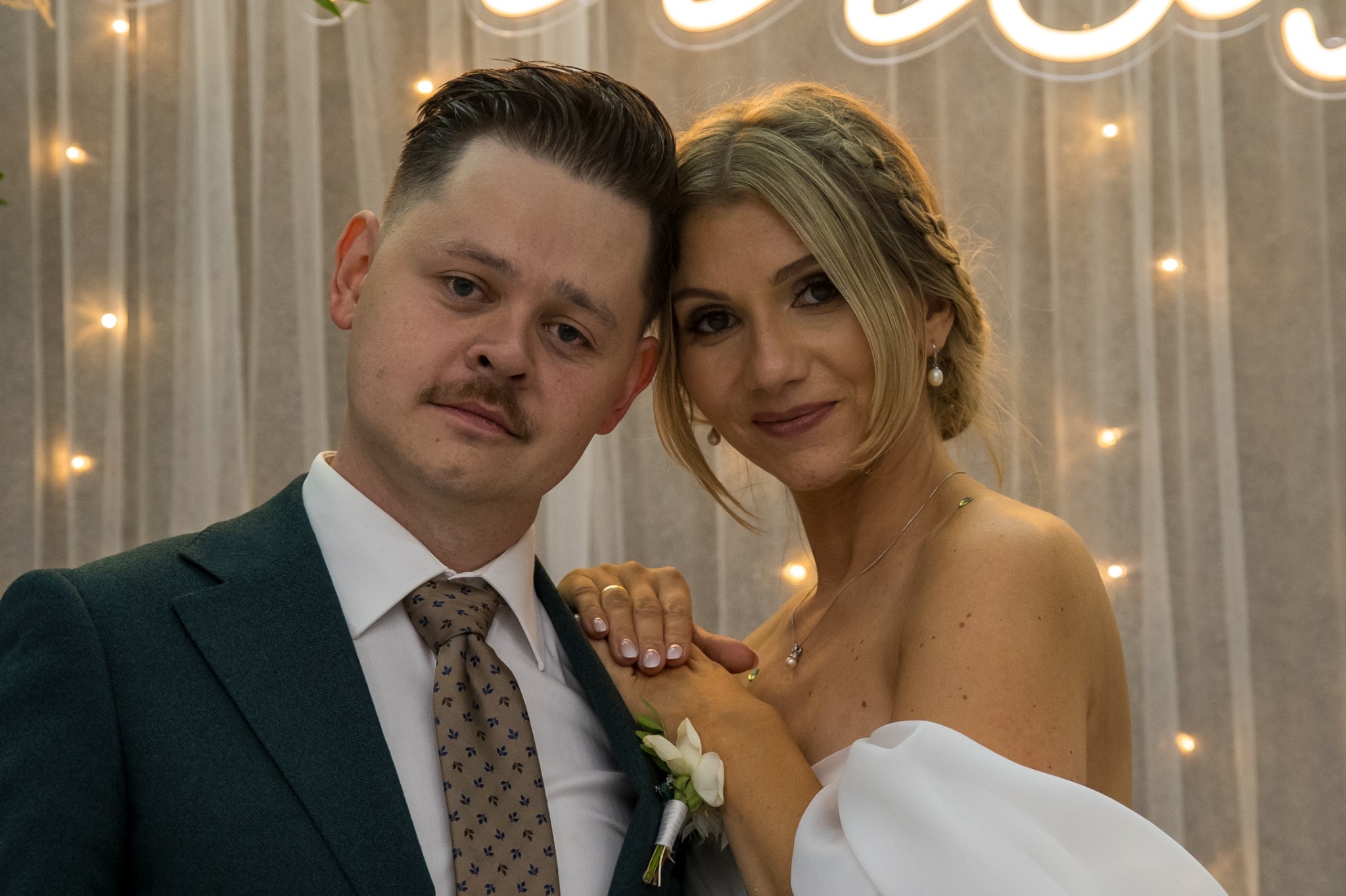 Portret pary młodej w zbliżeniu, on w ciemnozielonym garniturze, ona w białej sukni, na tle białej zasłony z delikatnymi światełkami, z subtelnym uśmiechem.