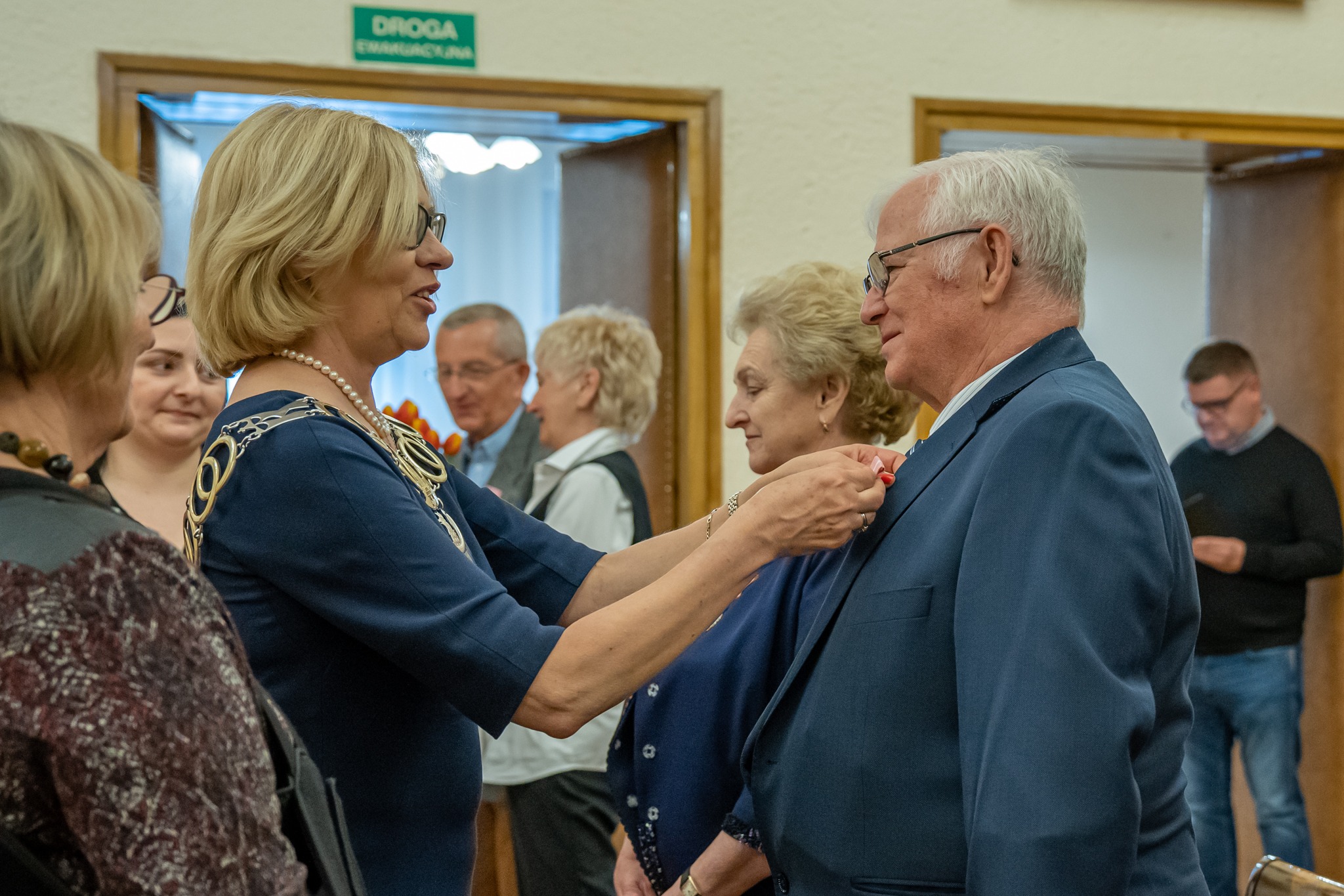 Uroczyste przypięcie odznaczenia mężczyźnie w garniturze przez kobietę w sukience, w tle zgromadzeni ludzie i tabliczka 'Droga Ewakuacyjna'.