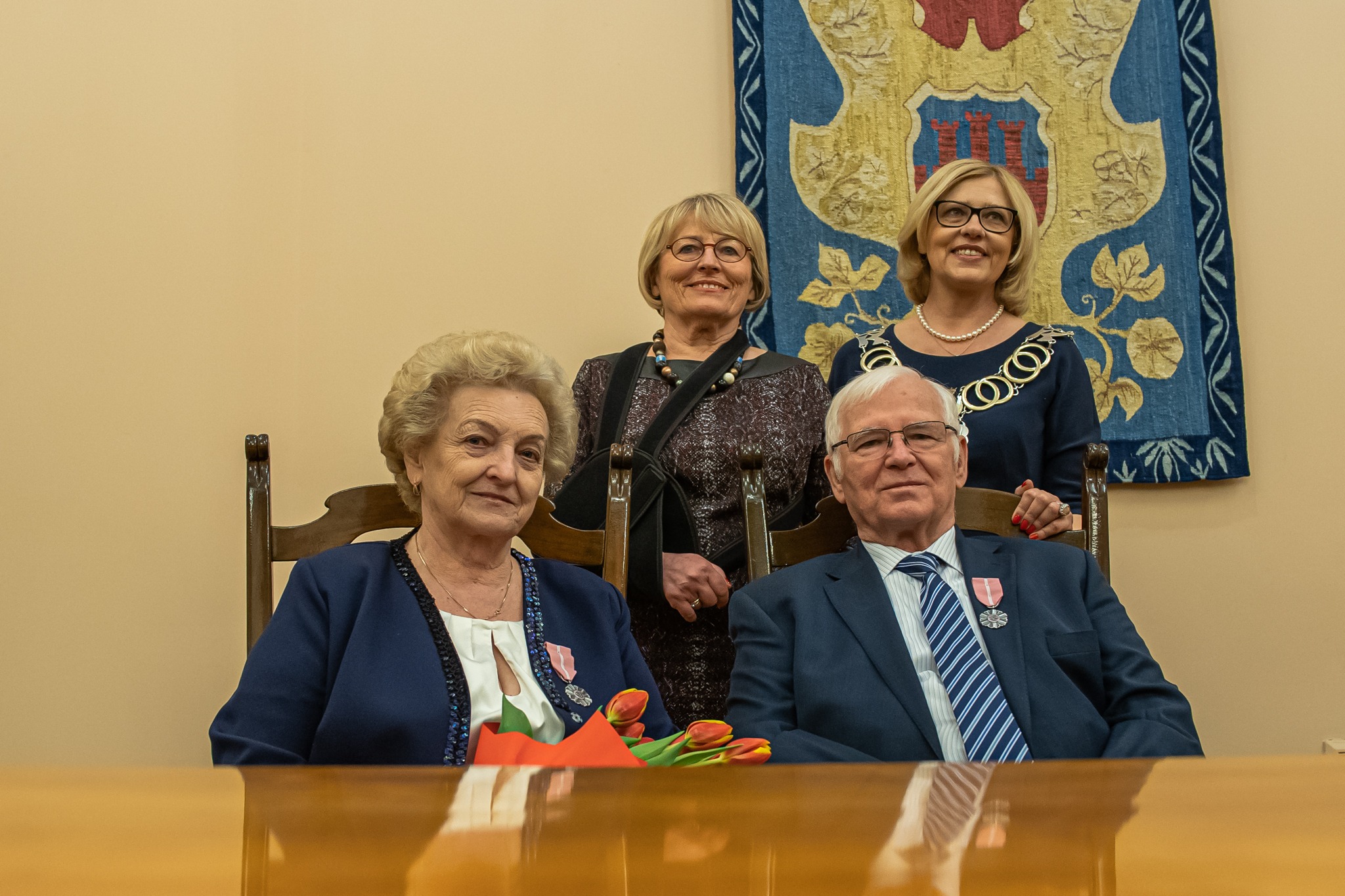 Uroczyste wręczenie odznaczeń państwowych: cztery osoby pozują do zdjęcia na tle herbu miasta. Dwie osoby siedzą, trzymają bukiet tulipanów.