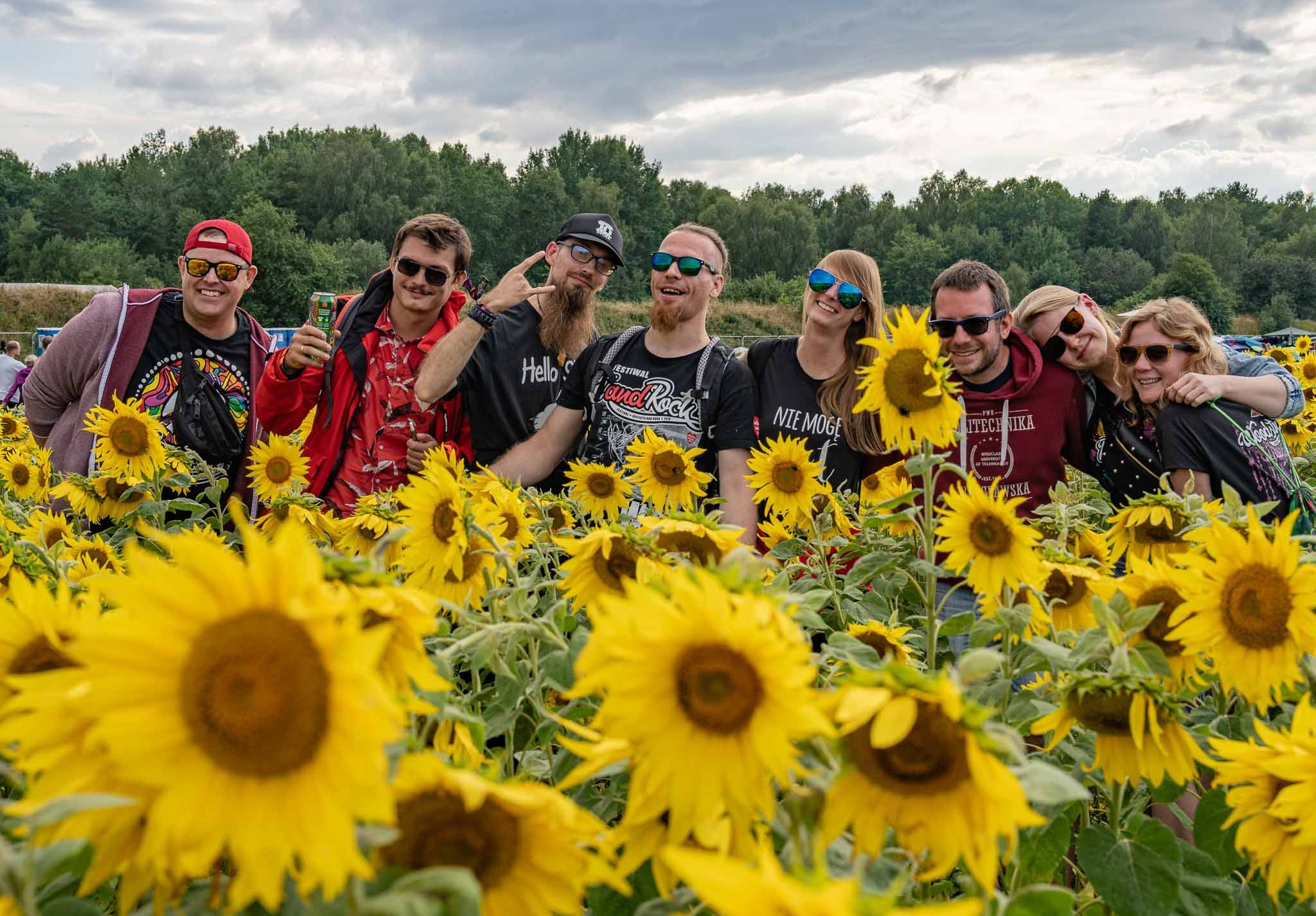 Grupa przyjaciół w okularach przeciwsłonecznych pozuje w polu słoneczników pod pochmurnym niebem. Jeden z mężczyzn trzyma puszkę napoju.