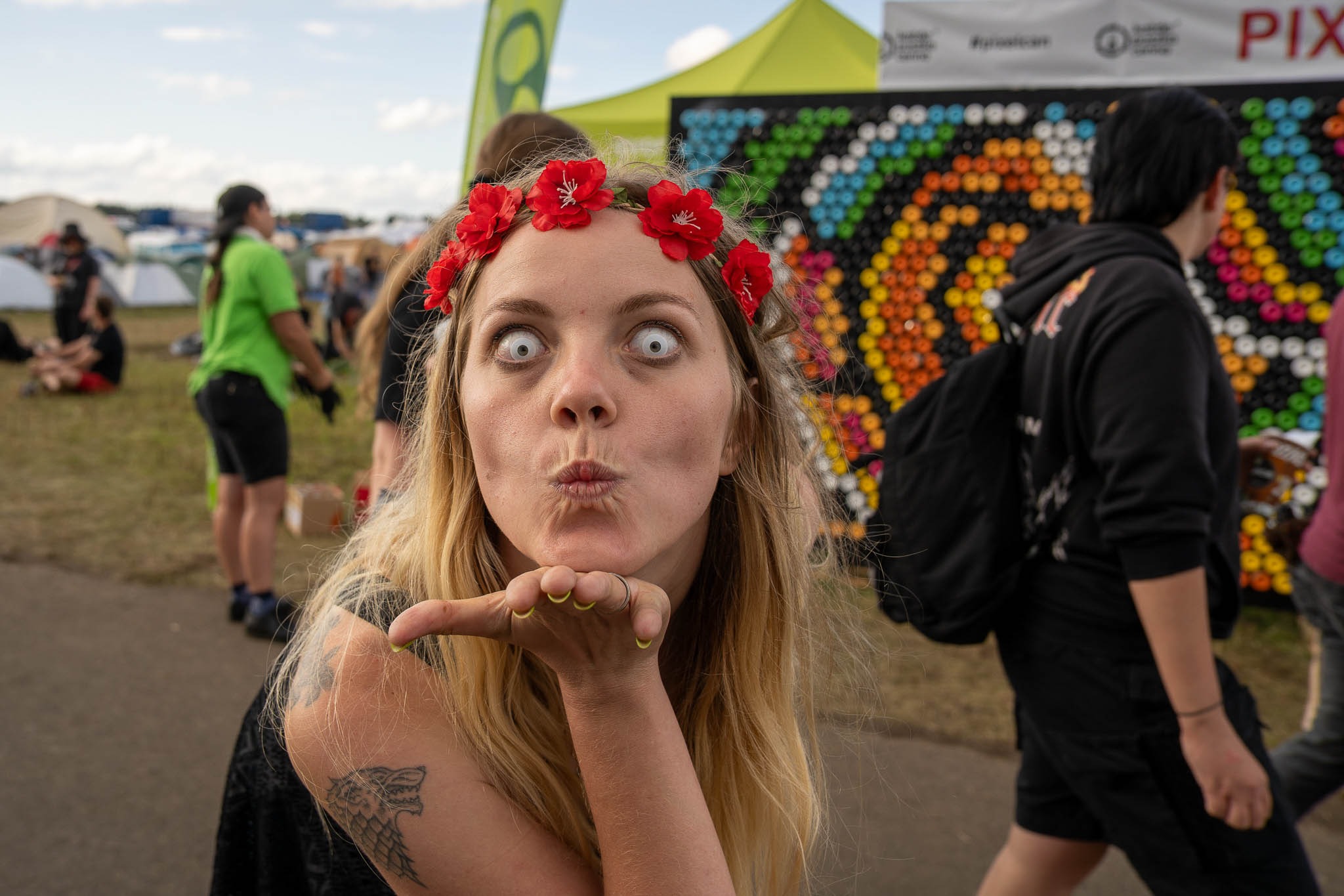 Portret kobiety z białymi soczewkami i wiankiem z czerwonych kwiatów na tle festiwalowym. Wyciągnięta dłoń, tatuaż wilka na ramieniu, ekspresyjna mimika.