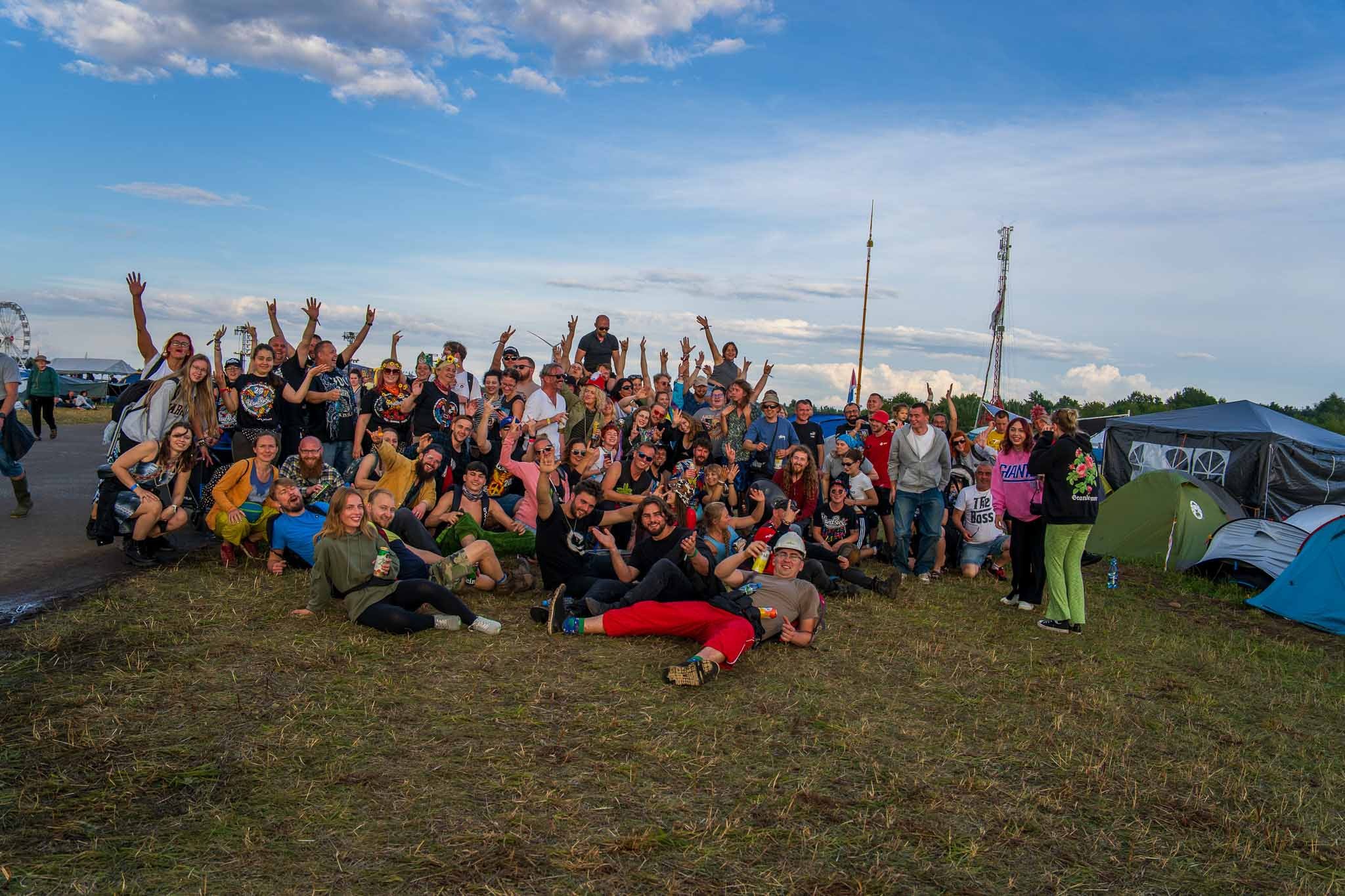 Duża grupa uśmiechniętych ludzi pozuje na tle pleneru z namiotami i konstrukcjami, unosząc ręce w geście radości podczas eventu.