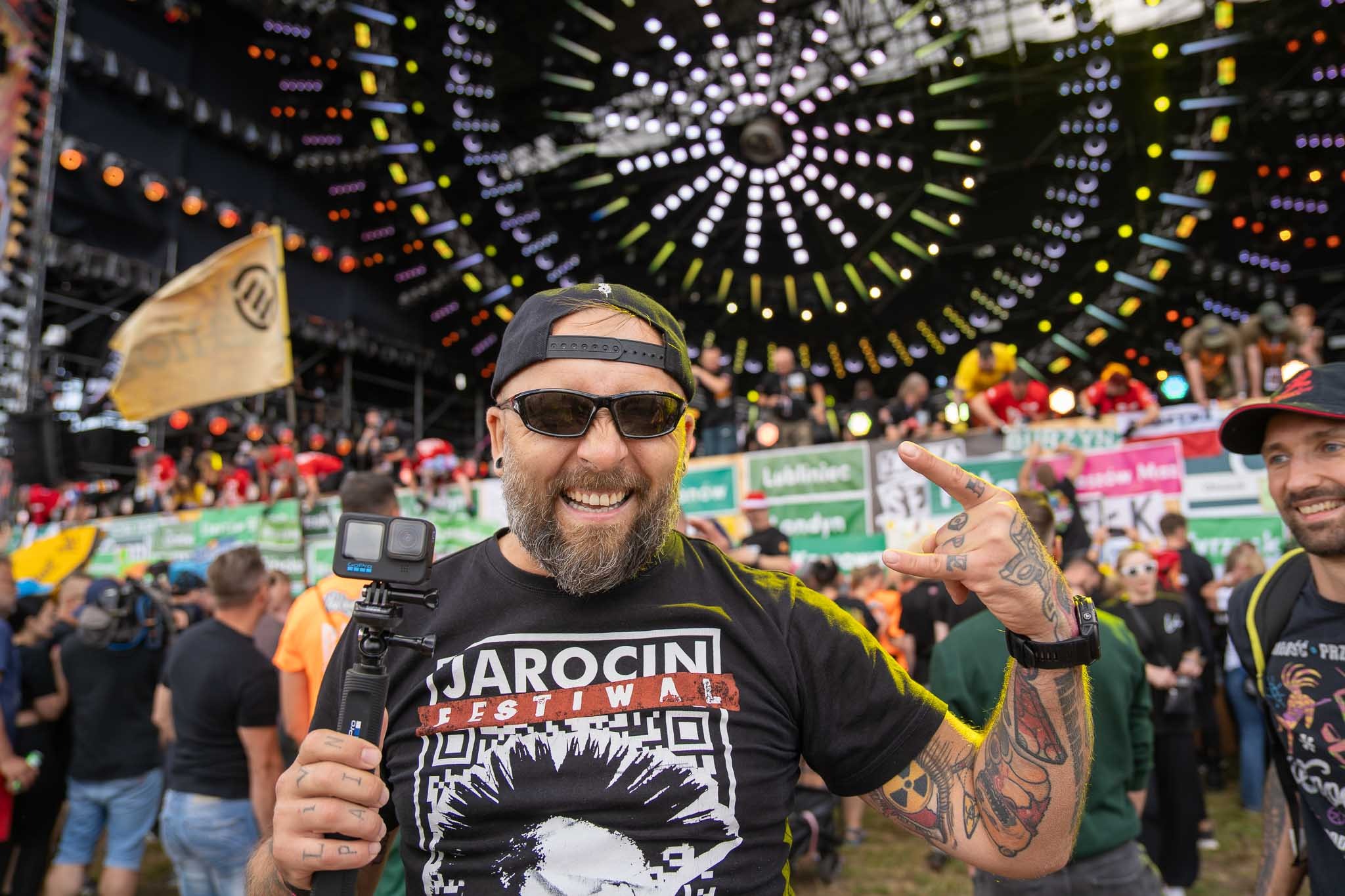Uśmiechnięty mężczyzna z brodą i tatuażami, w czarnej koszulce Jarocin Festival, trzyma kamerę GoPro na tle sceny z oświetleniem i publicznością.