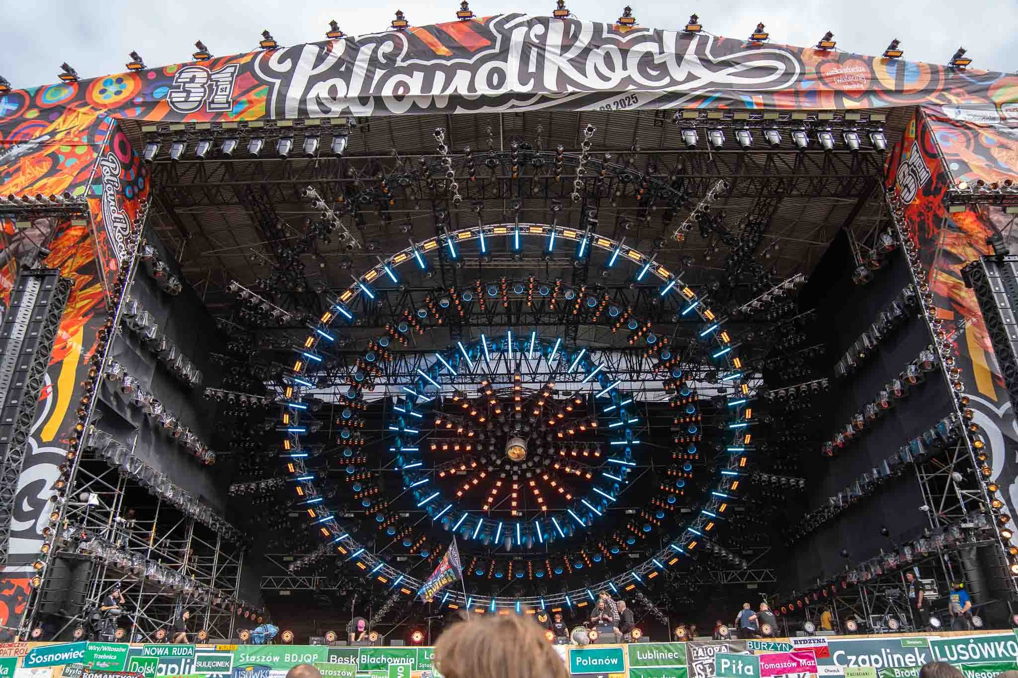 Scena koncertowa z rozbudowanym oświetleniem w kształcie okręgów, banerem 'PolandRock' i widoczną publicznością w dolnej części kadru. Scena przygotowana do występu.