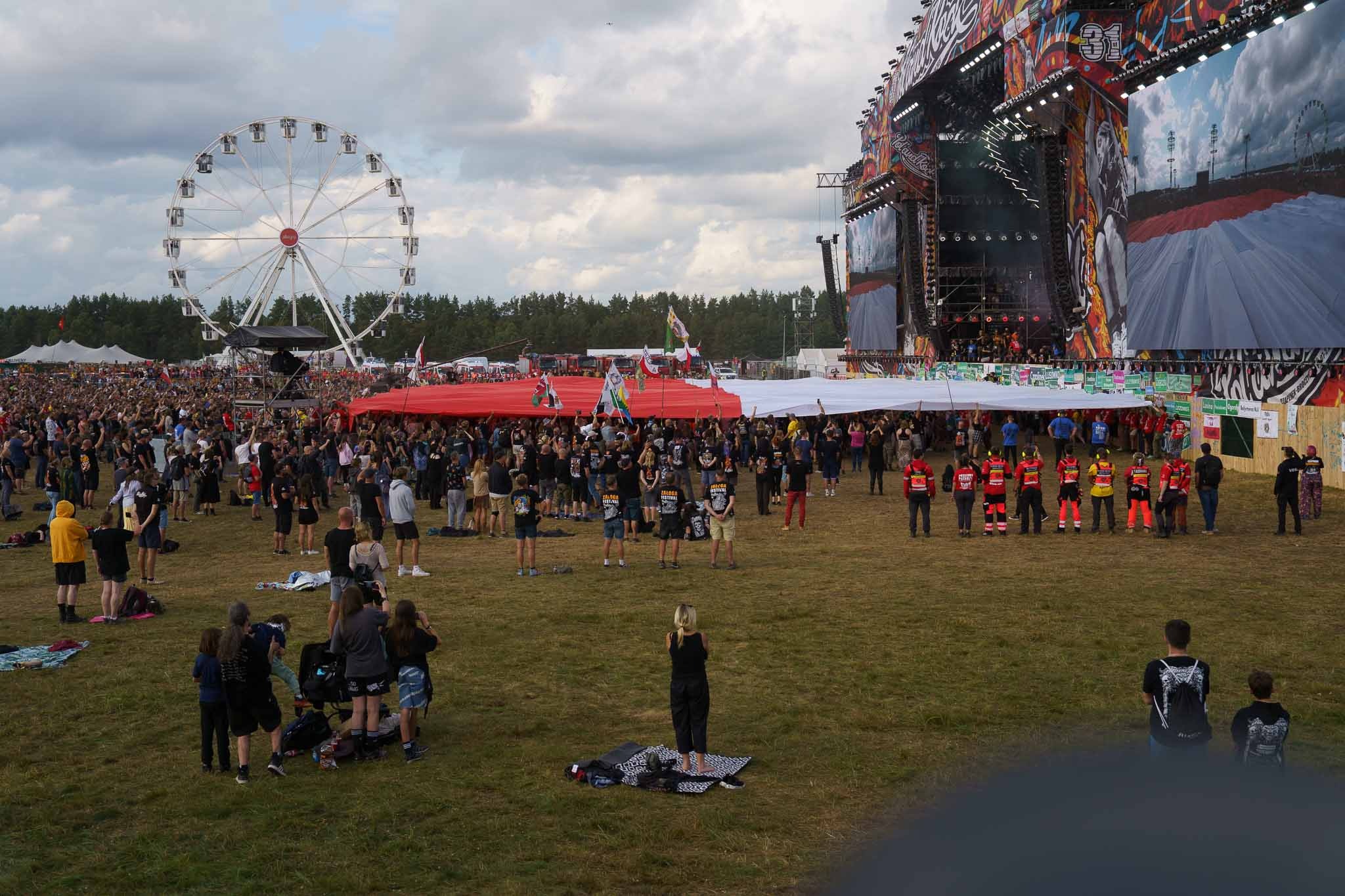 Tłum ludzi na festiwalu muzycznym z diabelskim młynem i sceną w tle. Uczestnicy trzymają duży, biało-czerwony transparent. Widok z perspektywy osoby stojącej na trawie.