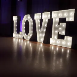 Podświetlany napis LOVE z żarówkami ustawiony na parkiecie, tło z fioletowym oświetleniem.