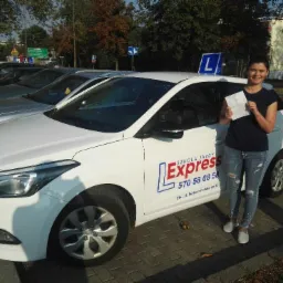 Uśmiechnięta kobieta trzymająca dokument, stojąca obok białego samochodu szkoły jazdy 'Express' z niebieską literą 'L' na dachu, na tle ulicy i drzew.