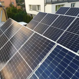 Instalacja fotowoltaiczna na płaskim dachu budynku mieszkalnego, widoczne moduły w układzie rzędowym, odbicia nieba na powierzchni paneli.