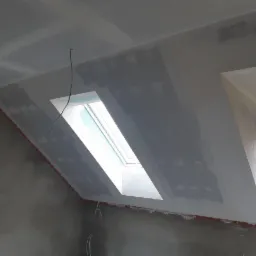 Poddasze w trakcie wykańczania: zamontowane płyty karton-gips, widoczne szpachlowanie łączeń i okno dachowe. W tle fragment ściany i okna.