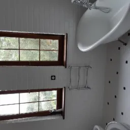 Biała łazienka z wanną na nóżkach, dwoma oknami z drewnianymi ramami, grzejnikiem drabinkowym i podłogą w biało-czarną szachownicę.