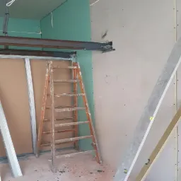 Montaż płyty karton gips: widok na drewnianą drabinę opartą o konstrukcję, profile metalowe, stalowy profil nad otworem, oraz poziomice opartą o ścianę z płyt gipsowych.