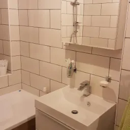 Nowoczesna łazienka z białymi, prostokątnymi płytkami, umywalką z szafką i kabiną prysznicową widoczną w lustrze.